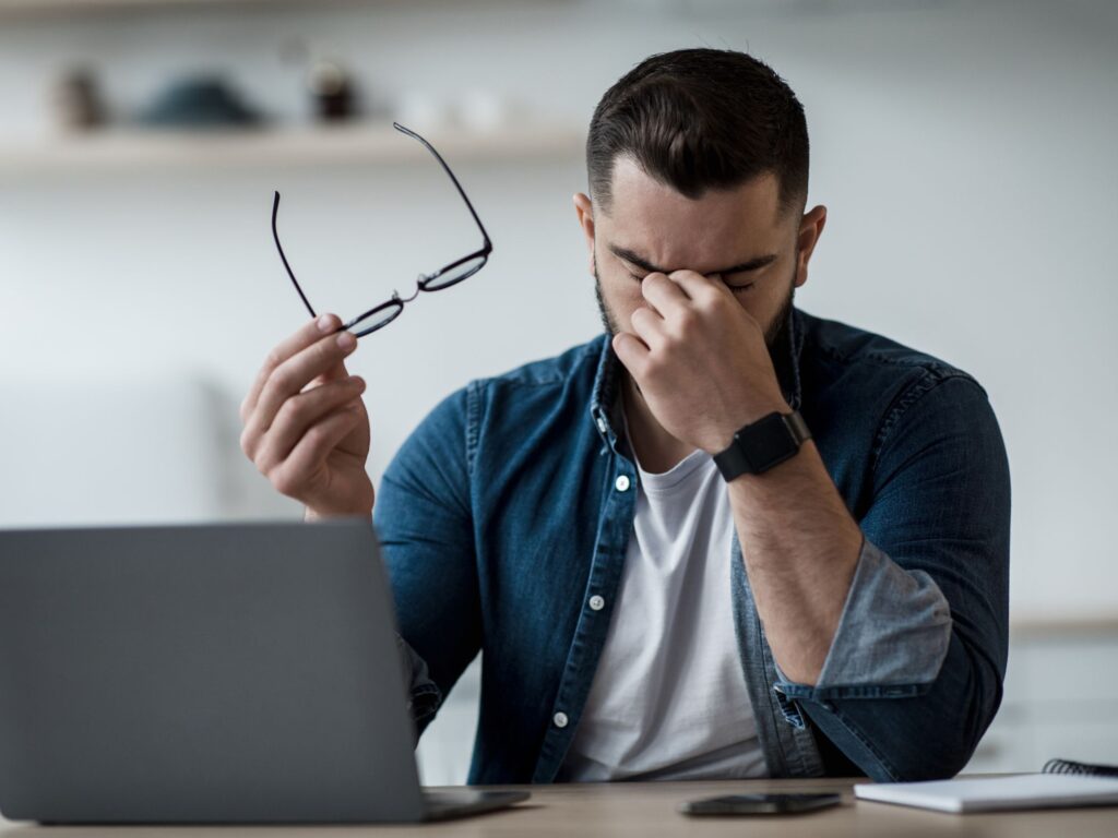 a man sitting in front of a laptop holding glasses in one hand and rubbing his eyes with the other hand, frowning